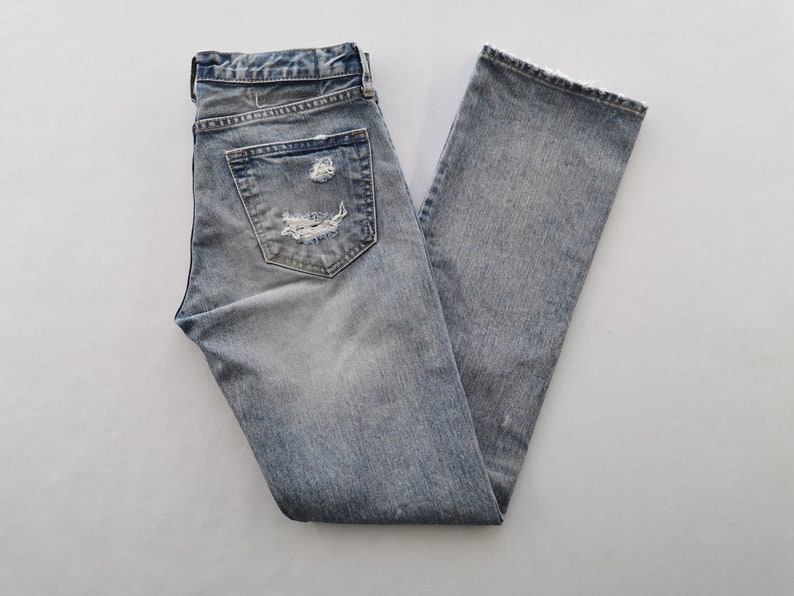 May include: Light blue denim jeans with distressed details, including a ripped pocket and a frayed hem. The jeans are folded and laid flat, showcasing their faded wash and classic five-pocket design.