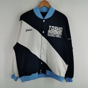 May include: A navy blue and white varsity jacket with light blue accents. The jacket features the text "TOSHO HIGHSCHOOL BASKETBALL" and the Asics logo. The jacket has a button-up front and is hanging on a wooden hanger.