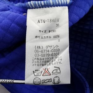 May include: Close-up of a blue garment with a white tag. The tag displays text including "ATN-1840W", size information, and care instructions. The fabric appears to be a textured polyester material.