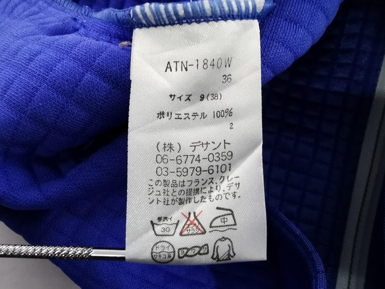 May include: Close-up of a blue garment with a white tag. The tag displays text including "ATN-1840W", size information, and care instructions. The fabric appears to be a textured polyester material.