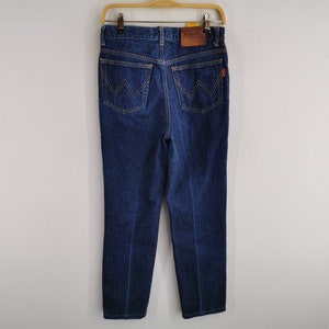 May include: Dark blue denim jeans hanging on a wooden hanger. The jeans have a classic five-pocket design with a leather patch and a red tag. The back pockets feature a distinctive 'W' stitching.