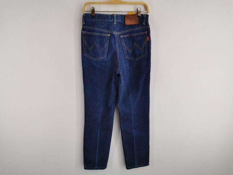 May include: Dark blue denim jeans hanging on a wooden hanger. The jeans have a classic five-pocket design with a leather patch and a red tag. The back pockets feature a distinctive 'W' stitching.
