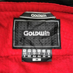 Pode incluir: Close-up de uma etiqueta preta com o nome da marca "GOLDWIN" em branco, sobre um tecido vermelho texturizado. A etiqueta inclui indicadores de tamanho e informa&ccedil;&otilde;es de tamanho internacionais.