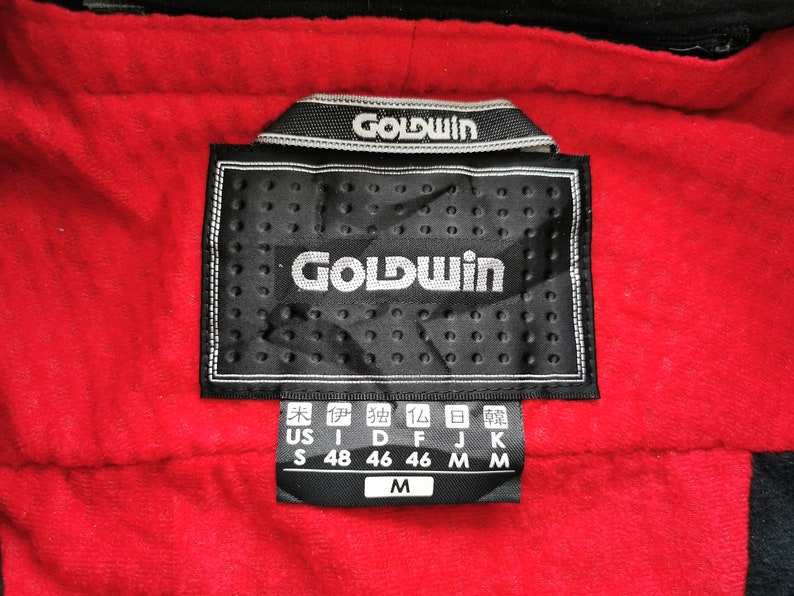 Pode incluir: Close-up de uma etiqueta preta com o nome da marca "GOLDWIN" em branco, sobre um tecido vermelho texturizado. A etiqueta inclui indicadores de tamanho e informa&ccedil;&otilde;es de tamanho internacionais.