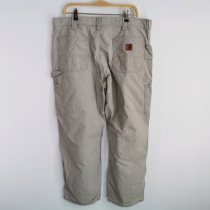 May include: Khaki-colored work pants with multiple pockets and a brown leather patch. The pants are hanging on a wooden hanger against a white wall. The pants have a relaxed fit and are made of a durable material.