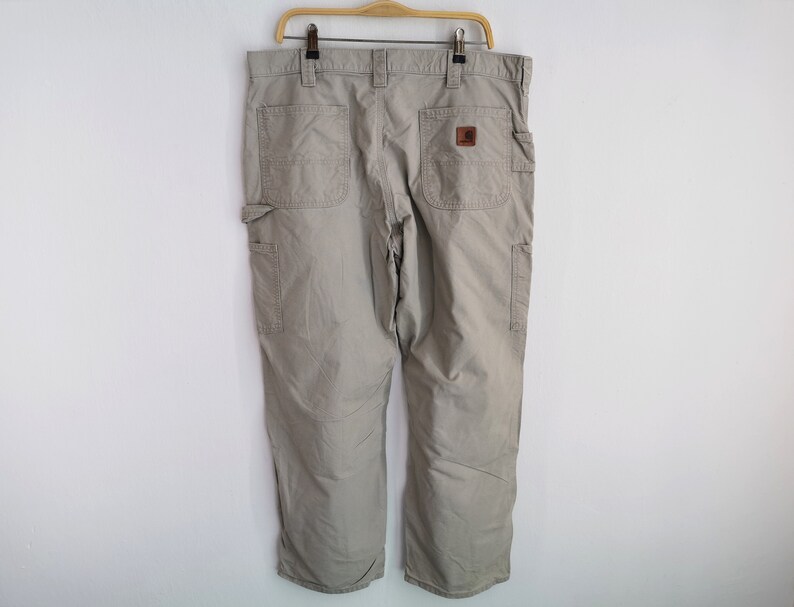 May include: Khaki-colored work pants with multiple pockets and a brown leather patch. The pants are hanging on a wooden hanger against a white wall. The pants have a relaxed fit and are made of a durable material.
