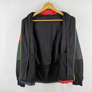 May include: Black zip-up jacket with a red interior and accents of green, gray, and blue. The jacket has a quilted pattern on the sleeves and is hanging on a wooden hanger against a white wall. The jacket is open, revealing the interior.