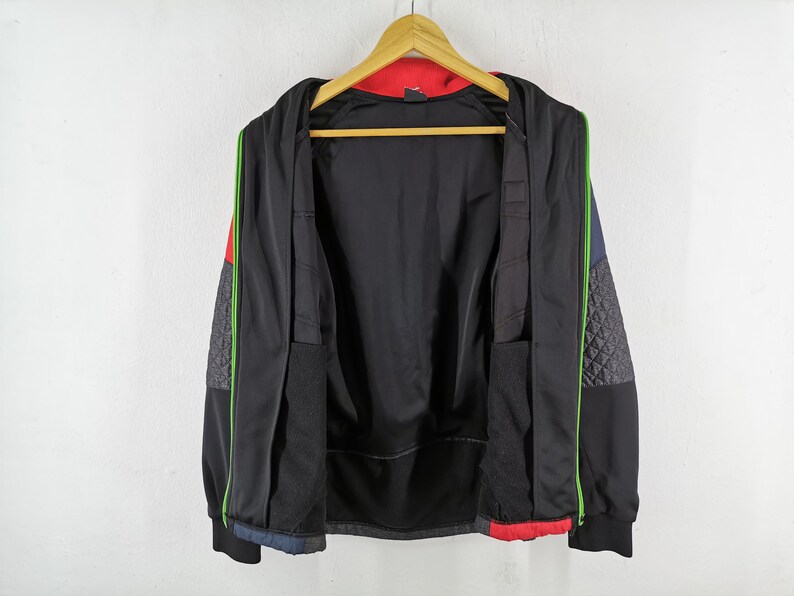 May include: Black zip-up jacket with a red interior and accents of green, gray, and blue. The jacket has a quilted pattern on the sleeves and is hanging on a wooden hanger against a white wall. The jacket is open, revealing the interior.