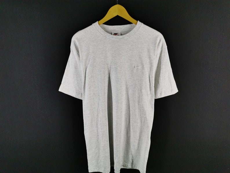 May include: A heather gray t-shirt with short sleeves, hanging on a wooden hanger. The shirt features a small Nike logo on the upper left chest. The background is a dark gray.