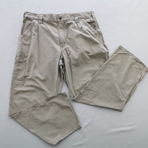 May include: Khaki-colored work pants with multiple pockets and a relaxed fit. The pants are made of a durable fabric and feature a button and zipper closure. The pants are laid flat on a light gray surface.
