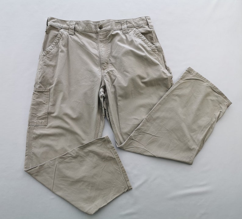 May include: Khaki-colored work pants with multiple pockets and a relaxed fit. The pants are made of a durable fabric and feature a button and zipper closure. The pants are laid flat on a light gray surface.
