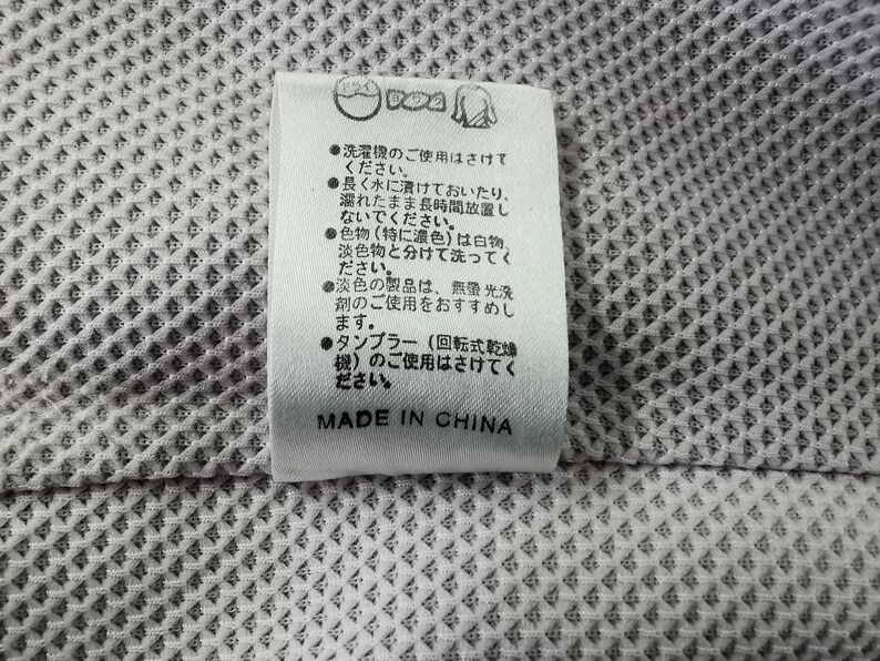 Puede incluir: Una etiqueta blanca con texto e iconos japoneses, adherida a una tela gris claro texturizada. La etiqueta incluye instrucciones de lavado y el texto "MADE IN CHINA".