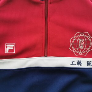 Puede incluir: Una chaqueta deportiva de cremallera corta en rojo, blanco y azul con un logotipo Fila blanco en el pecho derecho, un emblema bordado blanco en el pecho izquierdo y caracteres japoneses bordados en azul sobre la franja blanca del pecho.