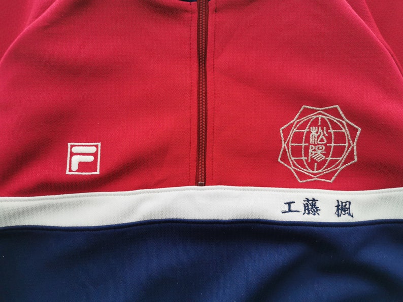 Puede incluir: Una chaqueta deportiva de cremallera corta en rojo, blanco y azul con un logotipo Fila blanco en el pecho derecho, un emblema bordado blanco en el pecho izquierdo y caracteres japoneses bordados en azul sobre la franja blanca del pecho.
