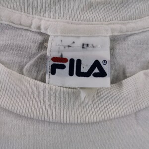 May include: Close-up of a white t-shirt collar with a Fila brand tag. The tag features the Fila logo in navy blue and red. The shirt's collar is ribbed and the fabric appears to be a soft cotton.