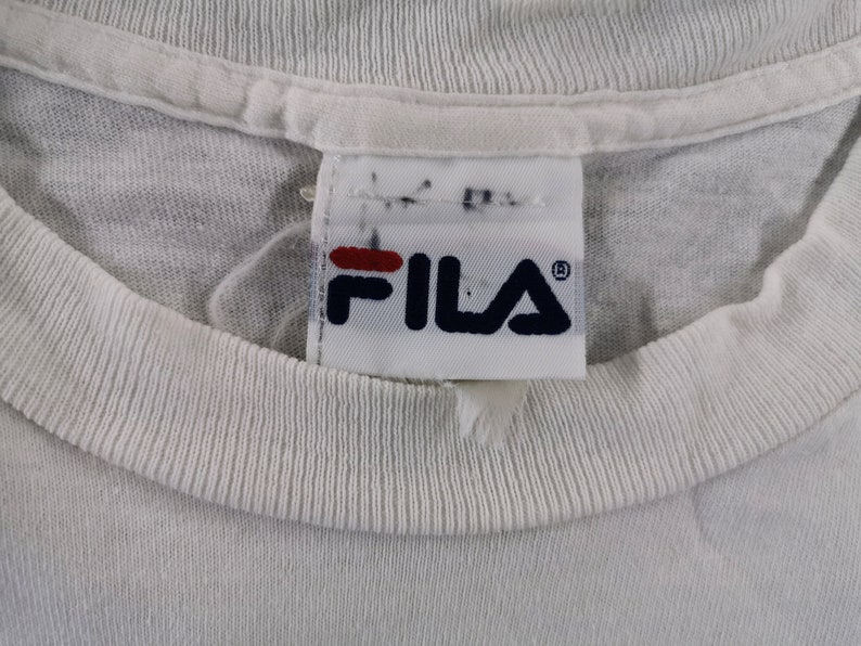 May include: Close-up of a white t-shirt collar with a Fila brand tag. The tag features the Fila logo in navy blue and red. The shirt's collar is ribbed and the fabric appears to be a soft cotton.