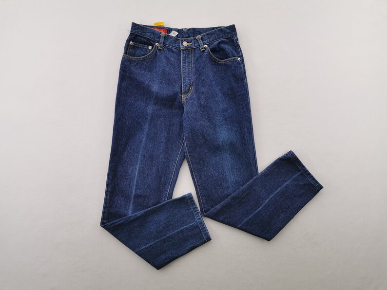 May include: Dark blue denim jeans with a classic five-pocket design. The jeans have a straight leg cut and a button and zipper closure. The stitching is a contrasting yellow color. The jeans are laid flat on a light gray surface.