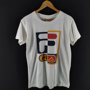 May include: White t-shirt with a large, colorful Fila logo. The logo is divided into sections of navy blue, red, and yellow. The shirt is hanging on a wooden hanger against a dark background.
