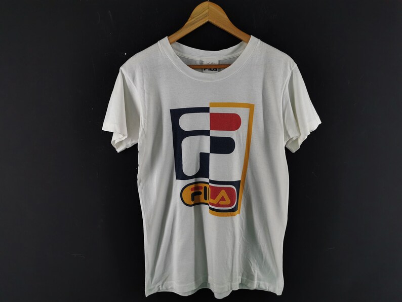 May include: White t-shirt with a large, colorful Fila logo. The logo is divided into sections of navy blue, red, and yellow. The shirt is hanging on a wooden hanger against a dark background.