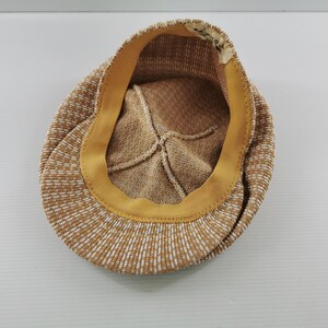 Puede incluir: Gorra de estilo newsboy con estampado marr&oacute;n y blanco. El interior presenta un forro beige y una banda amarilla. La gorra est&aacute; hecha de una tela texturizada. La visera no es visible.