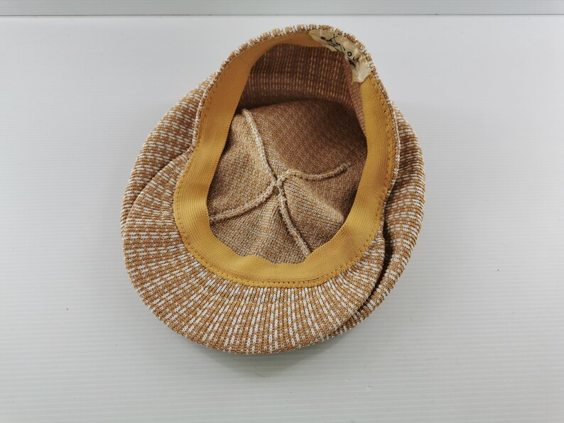 Puede incluir: Gorra de estilo newsboy con estampado marr&oacute;n y blanco. El interior presenta un forro beige y una banda amarilla. La gorra est&aacute; hecha de una tela texturizada. La visera no es visible.