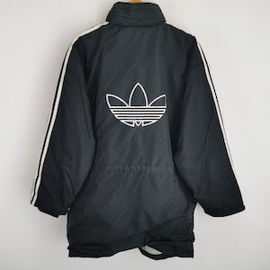 Puede incluir: Chaqueta Adidas negra con rayas blancas en las mangas y un gran logotipo blanco de Adidas en la espalda. La chaqueta tiene un cuello alto y una cintura ajustable. Una prenda deportiva clásica.