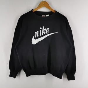 K&ouml;nnte beinhalten: Schwarzes Sweatshirt mit dem wei&szlig;en Nike-Logo auf der Brust. Das Logo zeigt den Markennamen in einer stilisierten Schriftart mit einem geschwungenen Haken. Das Sweatshirt hat einen Rundhalsausschnitt und lange &Auml;rmel.