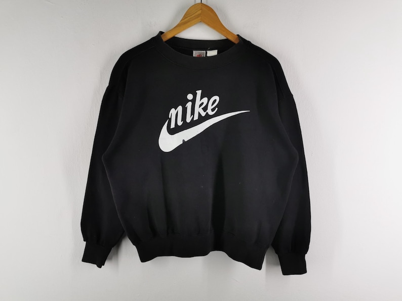 K&ouml;nnte beinhalten: Schwarzes Sweatshirt mit dem wei&szlig;en Nike-Logo auf der Brust. Das Logo zeigt den Markennamen in einer stilisierten Schriftart mit einem geschwungenen Haken. Das Sweatshirt hat einen Rundhalsausschnitt und lange &Auml;rmel.