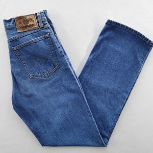 May include: Blue denim jeans with a classic five-pocket design. The jeans feature a straight leg cut and a brown leather patch with the "Wrangler" logo on the back. The jeans are folded neatly, showcasing the back pockets and the brand label.