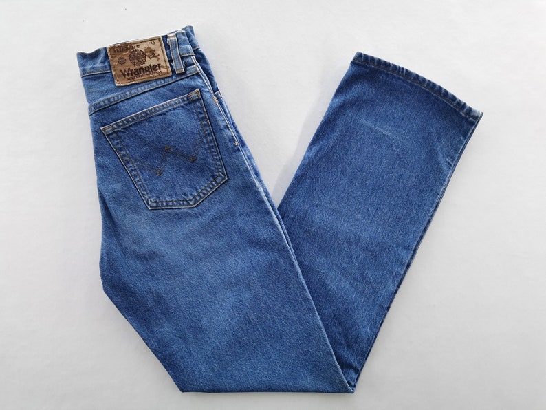 May include: Blue denim jeans with a classic five-pocket design. The jeans feature a straight leg cut and a brown leather patch with the "Wrangler" logo on the back. The jeans are folded neatly, showcasing the back pockets and the brand label.
