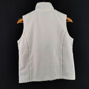 Puede incluir: Un chaleco blanco con cuello alto, exhibido en una percha de madera sobre un fondo negro. El chaleco tiene una apariencia acolchada y presenta costuras verticales en los laterales.