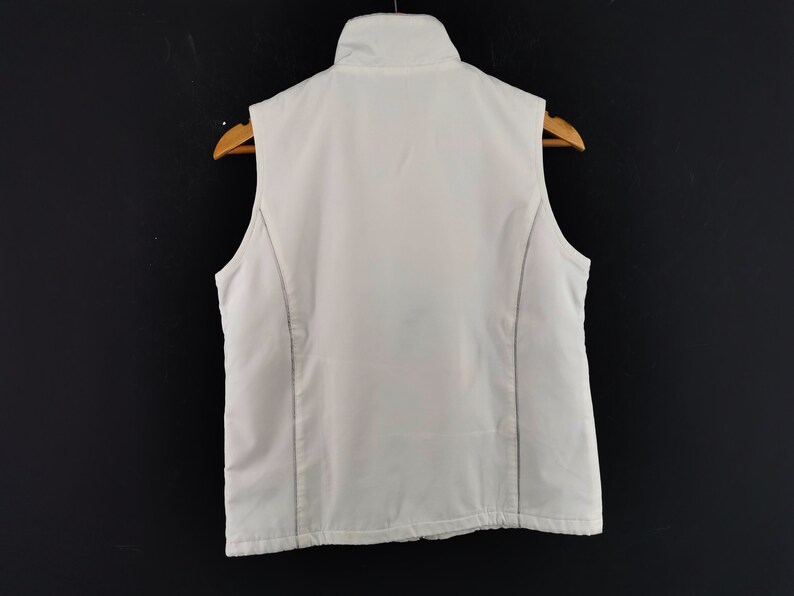 Puede incluir: Un chaleco blanco con cuello alto, exhibido en una percha de madera sobre un fondo negro. El chaleco tiene una apariencia acolchada y presenta costuras verticales en los laterales.