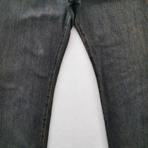 May include: Close-up of faded black denim jeans. The jeans have a classic five-pocket design and a straight leg cut. The stitching is a contrasting yellow-gold colour.