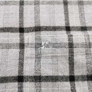 May include: Close-up of a gray and black plaid fabric. The fabric has a classic plaid pattern with intersecting lines in black and gray. A small pocket is visible, and there is a small tear in the fabric.