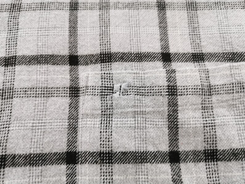 May include: Close-up of a gray and black plaid fabric. The fabric has a classic plaid pattern with intersecting lines in black and gray. A small pocket is visible, and there is a small tear in the fabric.