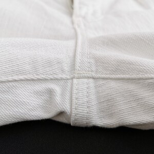 K&ouml;nnte beinhalten: Nahaufnahme von wei&szlig;em Denim-Stoff, der die Textur und die Nahtdetails zeigt. Das Bild hebt die N&auml;hte und das Gewebe des Materials hervor und deutet auf einen Fokus auf Kleidung oder Textilprodukte hin.