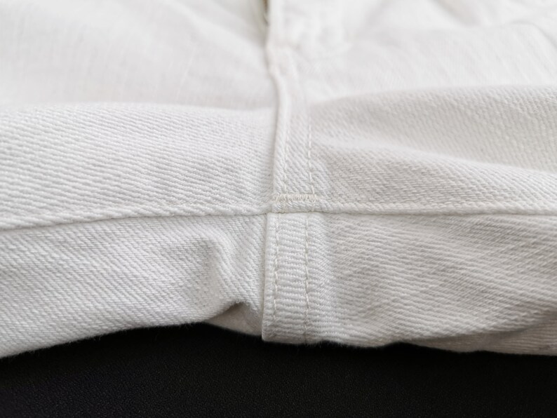 K&ouml;nnte beinhalten: Nahaufnahme von wei&szlig;em Denim-Stoff, der die Textur und die Nahtdetails zeigt. Das Bild hebt die N&auml;hte und das Gewebe des Materials hervor und deutet auf einen Fokus auf Kleidung oder Textilprodukte hin.