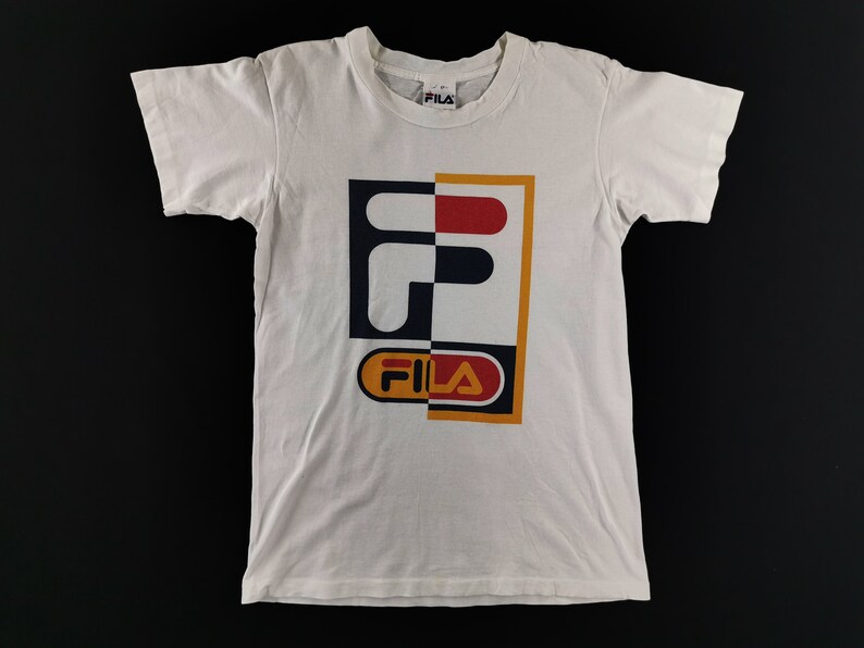 May include: White t-shirt with a large, colorful FILA logo on the front. The logo is split into two sections, with a combination of red, navy, yellow, and orange. The shirt has short sleeves and a crew neck.