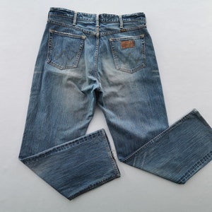 May include: Light blue denim jeans with a faded wash. The jeans feature classic five-pocket styling, a brown leather patch, and the Wrangler logo. The back pockets have a signature W-shaped stitching.