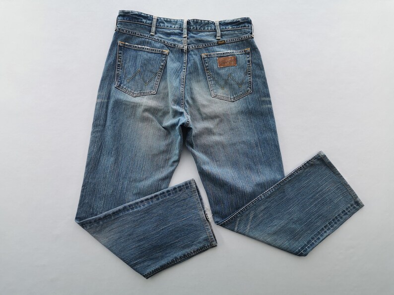 May include: Light blue denim jeans with a faded wash. The jeans feature classic five-pocket styling, a brown leather patch, and the Wrangler logo. The back pockets have a signature W-shaped stitching.