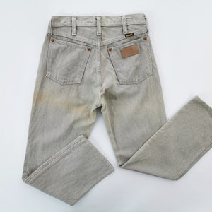 May include: Light grey Wrangler jeans with a faded appearance. The jeans have two back pockets, a small brown leather patch, and the Wrangler logo. The jeans are laid flat on a white surface.