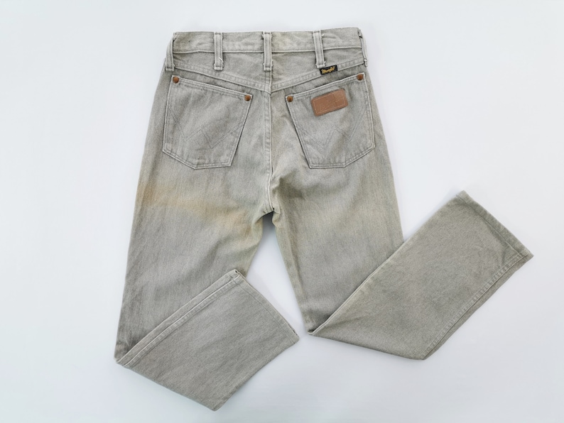 May include: Light grey Wrangler jeans with a faded appearance. The jeans have two back pockets, a small brown leather patch, and the Wrangler logo. The jeans are laid flat on a white surface.