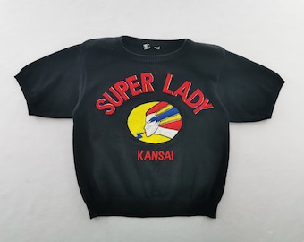 Puede incluir: Un suéter de punto negro de manga corta con el texto rojo SUPER LADY arqueado sobre un gráfico ovalado amarillo que contiene un perfil estilizado con cabello azul y rojo, con el texto rojo KANSAI impreso debajo del gráfico ovalado.