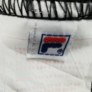 May include: Close-up of a white tag with the Fila logo in red, navy blue, and white. The tag is attached to black fabric and includes Korean text. The logo is a stylized "F" with a small emblem.