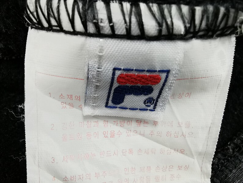 May include: Close-up of a white tag with the Fila logo in red, navy blue, and white. The tag is attached to black fabric and includes Korean text. The logo is a stylized "F" with a small emblem.