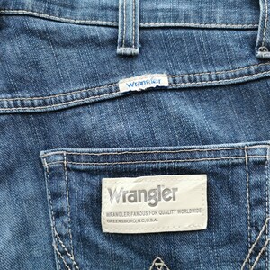 May include: Close-up of blue denim jeans with white stitching. The Wrangler brand label is visible on the back pocket, with the brand name and the text "Wrangler Famous for Quality Worldwide Greensboro, N.C., U.S.A."