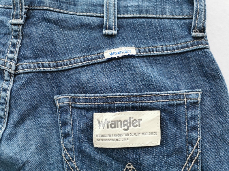 May include: Close-up of blue denim jeans with white stitching. The Wrangler brand label is visible on the back pocket, with the brand name and the text "Wrangler Famous for Quality Worldwide Greensboro, N.C., U.S.A."