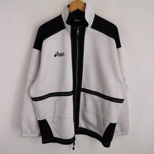 Puede incluir: Chaqueta con cremallera Asics gris claro y negra. La chaqueta presenta paneles negros en los hombros y a lo largo de la cremallera, con el logotipo de Asics bordado en el pecho. Tiene dos bolsillos delanteros y un cordón en la parte inferior.