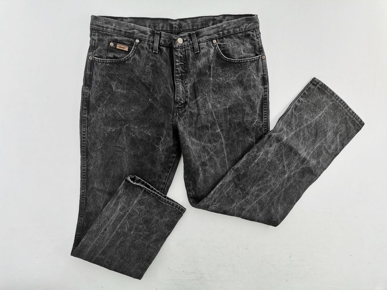May include: Acid wash black denim jeans. The jeans have a classic five-pocket design, a button and zipper closure, and a small logo patch on the front pocket. The denim has a faded, distressed look.