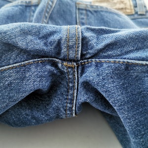 May include: Close-up of blue denim jeans, showcasing the stitching details at the crotch and waistband. The denim has a classic, worn look with visible texture and subtle fading. The stitching is in a contrasting tan color.